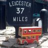 Red wooden double decker bus playset in situ sat infront of sign reading 'Leicester 37 miles'
